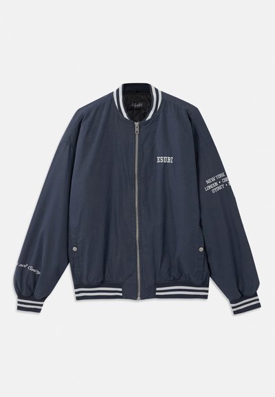 Jūras spēku bumbas jaka ar baltām svītrām, ar rāvējslēdzēju un izšūtu tekstu uz krūtīm un piedurknēm. Izgatavota no gludas auduma.