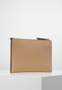 Bežová obdélníková kožená kabelka s texturovaným povrchem, zipovým zavíráním a střapcem, umístěná na bílém podstavci na jednolitém pozadí.