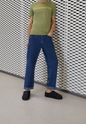 Mężczyzna w oliwkowozielonej koszulce Calvin Klein Jeans, niebieskich podwiniętych dżinsach i czarnych wsuwanych butach stoi na tle białej ściany z wzorem.