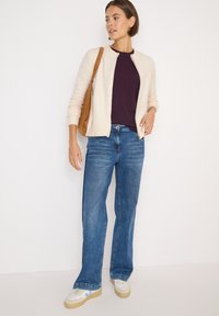 Beige stickad cardigan över en mörklila topp, ihop med ljusblå utsvängda jeans och vita och bruna sneakers. Brun axelväska.