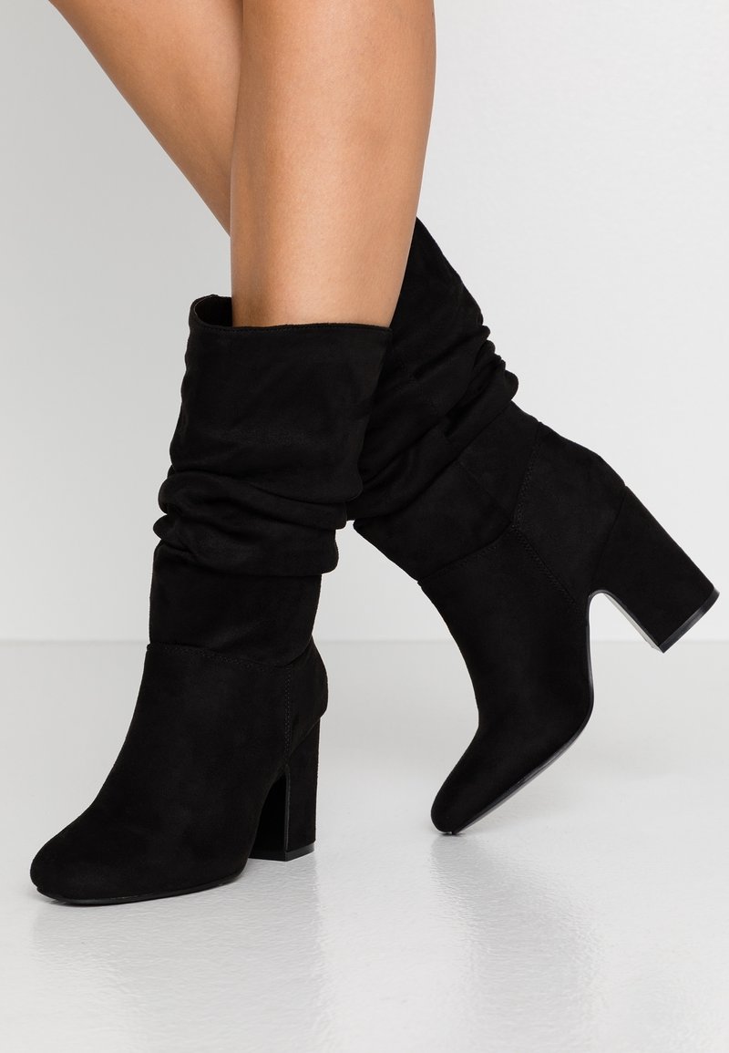 Mustat nahkaiset nilkkurit, joilla on löysä muotoilu, kapea kärki ja paksu korko, jotka erottuvat sileästä pinnastaan ja yksinkertaisesta rakenteestaan.