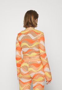 Top cropped em laranja, amarelo e creme com padrão ondulado. Mangas longas com um design ajustado. Textura de malha com um ligeiro brilho.