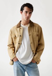 Jonge man in een beige jasje over een wit shirt en lichtblauwe spijkerbroek, staande met zijn handen in zijn zakken tegen een effen achtergrond.