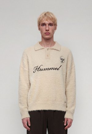 Jeune homme aux cheveux blonds courts portant un pull en molleton crème avec le texte "Hummel" et un petit logo d'abeille sur la poitrine, debout devant un fond uni.