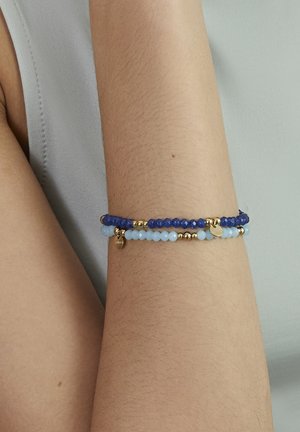 Arm wearing two beaded bracelets, one dark blue and one light blue with small gold-colored beads and round gold charms.