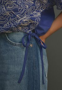 Modrá halenka s bílou abstraktní výšivkou na spodním lemu, kombinovaná s džínami s modrou stuhou v pase a detaily kapes.