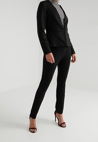 Blazer noir ajusté avec un décolleté en V, associé à un pantalon noir slim, et un haut rayé noir et blanc, porté avec des sandales à talons hauts.