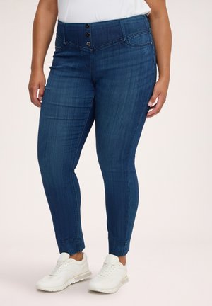Kvinde iført mørkeblå tætsiddende jeans med tre forreste knapper og hvide sneakers mod en ensfarvet baggrund.