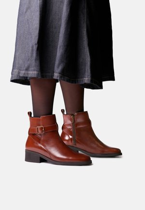 Bottines en cuir marron avec fermeture éclair latérale et boucle, portées avec des collants noirs et une jupe foncée.