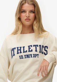 Sudadera color crema con texto y bordado en azul marino que dice "ATLETISMO". Corte relajado con puños acanalados y cuello redondo.