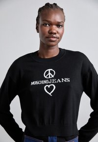 Melna adīta džempera, kas dekorēts ar baltu izšūtu mieru simbolu, tekstu "MOSCHINOJEANS" un sirds dizainu. Rievotas piedurknes un apakšmala.