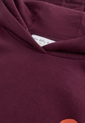 Bordová mikina s kapucňou, ktorá má mäkkú textúru, šnúrovú kapucňu a oranžový kruhový dizajnový akcent na prednej strane. Na vnútornej strane je čiastočne viditeľná etiketa.