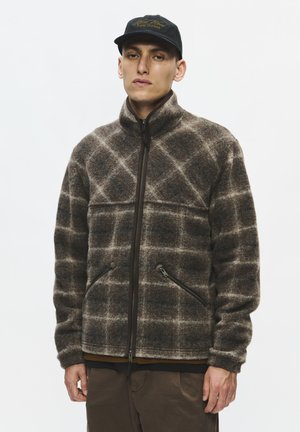 Homme portant une veste polaire à carreaux marron et beige avec des poches zippées et une casquette de baseball noire, debout devant un fond blanc uni.