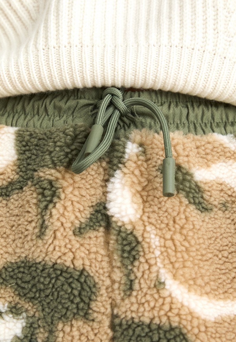 Szorty w wzór moro z pluszowego materiału w odcieniach zieleni i beżu. Posiadają sznurki z plastikowymi końcówkami oraz elastyczny pas.