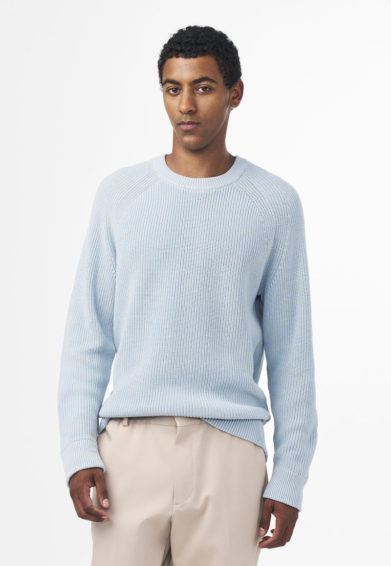 Maglione azzurro chiaro in lavorazione a maglia con texture a coste, collo rotondo e maniche lunghe, abbinato a pantaloni beige. È visibile un delicato motivo a righe blu.