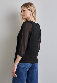 Blusa nera con maniche puff e trasparenti e schiena a chiave. Il tessuto ha una finitura testurizzata. Abbinata a jeans in denim blu a gamba larga.