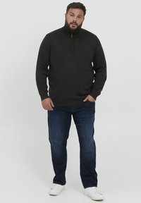 Suéter negro de cuello alto con cremallera y puños acanalados, combinado con jeans azul oscuro y zapatillas blancas. El conjunto presenta un diseño discreto y casual.