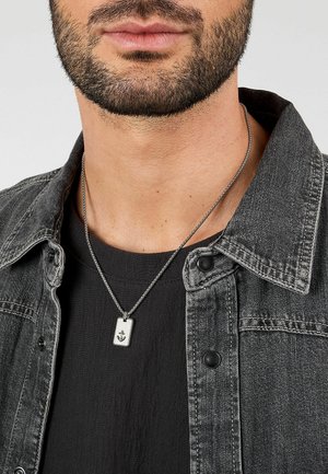 Collana con pendente d'argento con dettaglio di ancora, indossata da una persona che porta una giacca di jeans grigio scuro e una T-shirt nera. Vista ravvicinata.