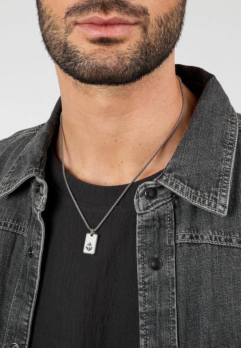 Collana con pendente d'argento con dettaglio di ancora, indossata da una persona che porta una giacca di jeans grigio scuro e una T-shirt nera. Vista ravvicinata.