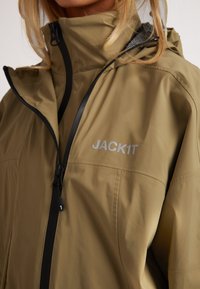 Giacca impermeabile verde oliva con colletto alto, zip nera e logo riflettente "JACK1T" sul petto. Texture liscia.