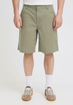 Uomo che indossa pantaloncini verde oliva, calzini bianchi alla caviglia e scarpe da ginnastica beige, in piedi davanti a uno sfondo semplice.