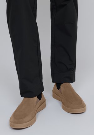 Hnědé semišové slip-on boty s tlustou béžovou podrážkou. Černé kalhoty zakrývají kotníky, což ukazuje na zaoblenou špičku boty a minimalistický design.