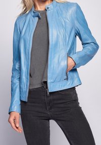 Kvinde iført en tætsiddende lyseblå læderjakke over en grå top og sorte jeans, med den ene hånd i jakkelommen.