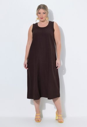 Kvinde iført sort, ærmeløs, løs midi-kjole og gule højhælede sandaler, stående foran en hvid baggrund.