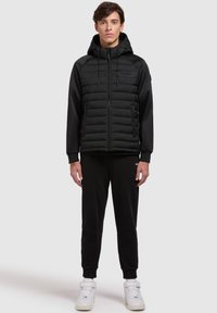 Zwarte gewatteerde jas met een capuchon en een voorzak, met een geplooide textuur, zijzakken en accentstiksels. Model draagt zwarte broek en witte sneakers.