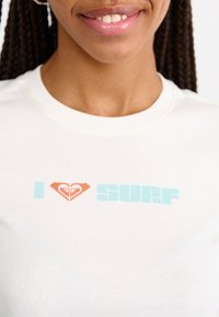 T-shirt en coton blanc comportant le texte "J ♥ SURF" en coeurs bleu clair et rouge, avec une texture lisse et un col rond.