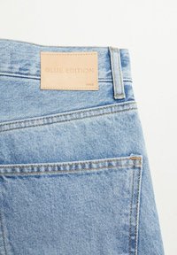 Vaaleansiniset farkut, joissa on sileä pinta, nahkainen merkki, jossa lukee "BLUE EDITION", ja näkyvät saumat taskujen kohdalla.