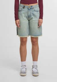 Persona vestida con pantalones cortos de mezclilla azul claro con los dobladillos deshilachados, una camiseta de manga larga color burdeos, calcetines blancos y zapatillas Adidas grises.