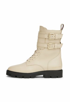 Botte de combat en cuir beige avec semelle à crampons noire, lacets à l'avant, deux sangles réglables à boucle, et languette de talon marquée.