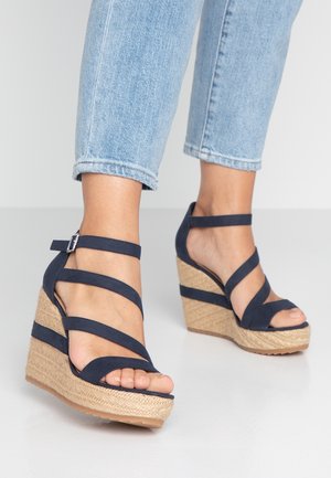 Stopy w granatowych sandałach na koturnie z paskami wokół kostki oraz jasno niebieskich, przyciętych dżinsach na jasnym, jednolitym tle.