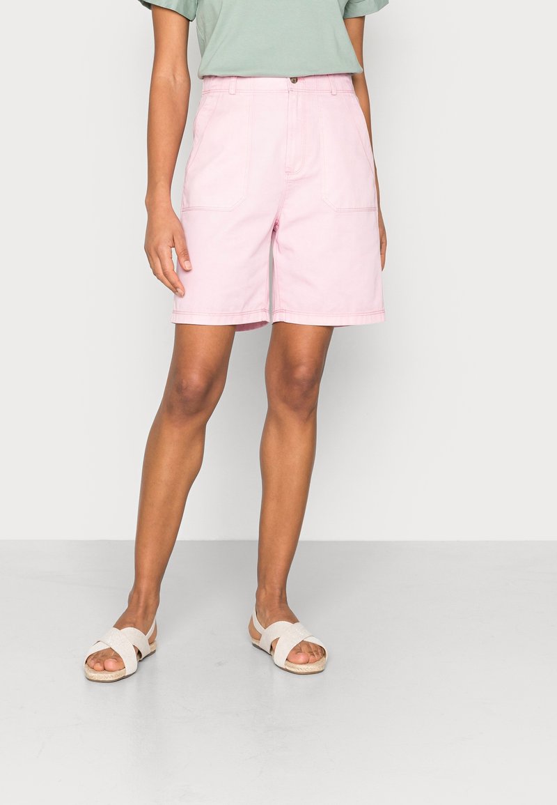 Persona che indossa pantaloncini rosa chiaro a vita alta con grandi tasche frontali, una camicia verde pastello infilata dentro e sandali beige con fasce incrociate.