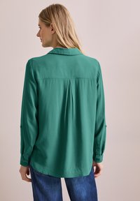 Blusa verde a maniche lunghe con una sottile texture, caratterizzata da una piega sul retro e polsini con bottoni, progettata con un colletto classico e una vestibilità morbida.