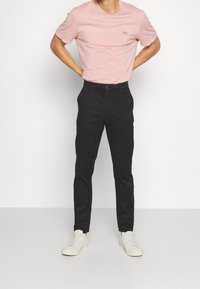 Ljusrosa kortärmad t-shirt med logotyp på bröstet, kombinerad med svarta slim-fit byxor och vita sneakers. Enkel design, mjuk textur.