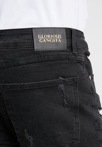 Jeans de mezclilla negra con textura desgastada, con una etiqueta con acentos dorados que dice "GLORIOUS GANGSTA" en la cintura y detalles de costura visibles.