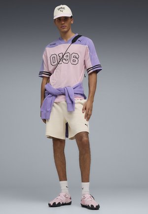 Giovane uomo che indossa una maglia sportiva rosa e viola, pantaloncini beige, scarpe da ginnastica rosa, cappellino bianco e una felpa viola legata in vita.