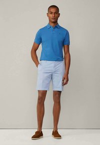 Modrá polo košile s krátkým rukávem v kombinaci se světle modrými šortkami. Oblečení zahrnuje hnědé boty na klínku. Jednoduchý design, pohodlný střih a měkké materiály.