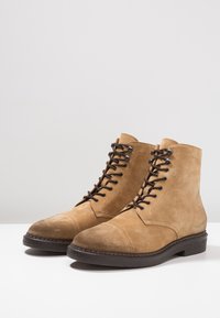 Un par de botas de ante color beige con cordones, con suelas marrón oscuro y cordones negros, exhibidas sobre una superficie blanca contra un fondo sencillo.
