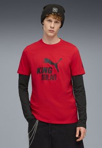 Rød T-shirt med korte ærmer, prydet med sort logo tekst "KING MILAN" og en sort Puma silhuet. Laget over en sort langærmet top.