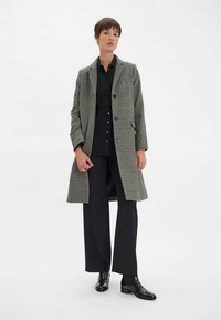 Manteau en laine à carreaux noir et blanc, à boutonnage simple avec des revers, associé à une chemise noire à boutons et un pantalon noir à jambes larges.