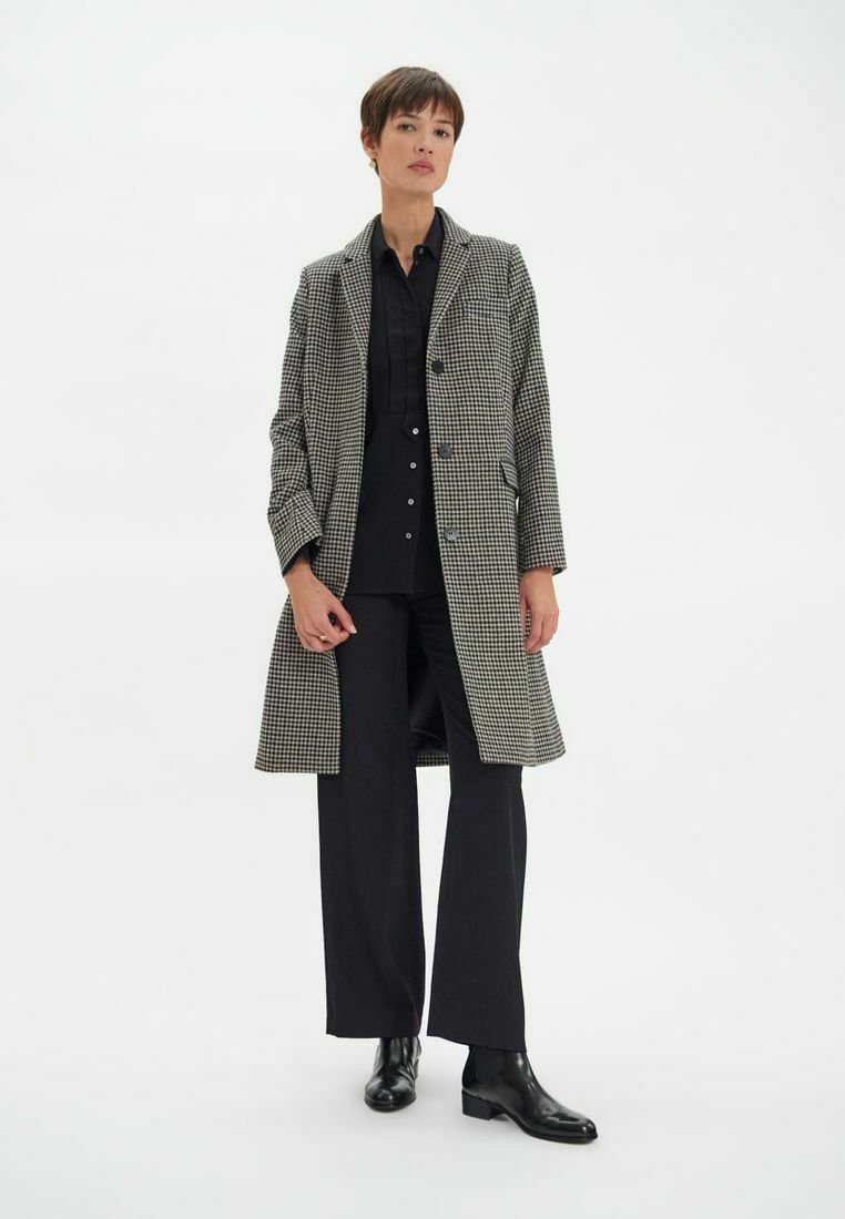 Manteau en laine à carreaux noir et blanc, à boutonnage simple avec des revers, associé à une chemise noire à boutons et un pantalon noir à jambes larges.