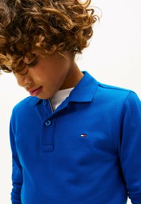 Polo bleu en tissu texturé, doté d'un rabat à deux boutons et d'un petit logo avec des accents rouge, blanc et bleu.