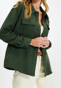 Camicia verde con bottoni, dal tessuto morbido, dotata di due tasche sul petto, maniche lunghe e colletto. Indossata sopra un top leggero a maniche corte.