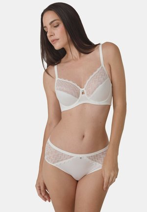 Woman wearing matching white lace bra and panties, standing with her head slightly tilted and eyes closed against a plain background.