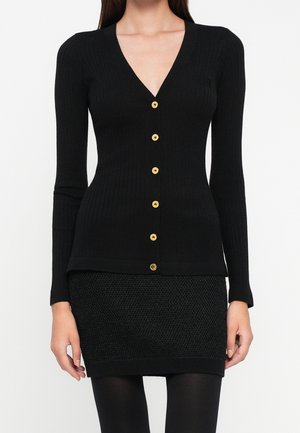 Kvinde iført en tætsiddende sort ribbet cardigan med guldknapper og et tekstureret sort mini nederdel, stående mod en hvid baggrund.