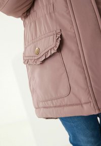 Gros plan sur la poche d'une veste matelassée mauve avec rabat à volants et bouton-pression doré, associée à un jean en denim bleu.