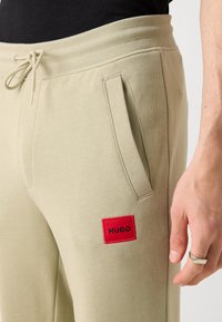 Beige sweatpants with a drawstring waist, side pockets, and a red square patch labeled "HUGO." Smooth texture and relaxed fit.